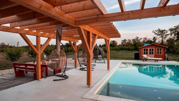 A wooden pergola with a pool, outdoor seating, and a small cabin in the background.