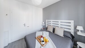 Un dormitorio moderno con una paleta de colores gris y blanco, una cama bien hecha, una mesita de noche con lámpara y una bandeja con artículos para el desayuno.