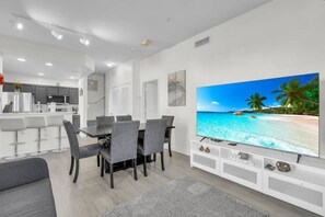 A modern living room with a dining area, a kitchen in the background, and a large flat-screen TV displaying a beach scene. - 4 Bedroom 3 Bathroom Villas At Seven Dwarfs 24-107 (Kissimmee)