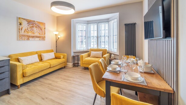 A modern living room with a sofa, dining table set, and a large window with blinds. - Modern 2 En-Suite Bedrooms Worcester, Free Parking (Worcestershire)