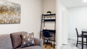 A living room with a grey sofa, a patterned pillow, a black office chair, a desk with a computer, and a bookshelf with plants.