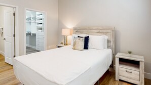 A bedroom with a large bed, a bedside table with a lamp, a wooden headboard, and a closet with hanging clothes.