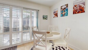 Un coin repas avec une table blanche, des chaises et une fenêtre équipée de stores.