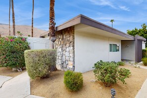 A modern house with a stone facade, a small front porch, and a well-maintained garden with various shrubs and a palm tree. - Beautiful Condo with mountain views, shared pool & pool spa (Palm Springs)
