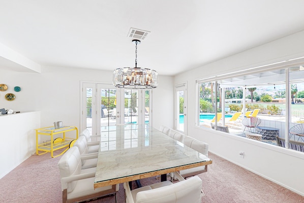 A spacious room with a large glass-top table, white chairs, and a view of a pool and outdoor seating. - Bright, Family-Friendly Home with Private Firepit, Heated Outdoor Pool, Pool Spa (Palm Springs)