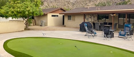 A residential setting with a small house, a patio with outdoor furniture, and a putting green.