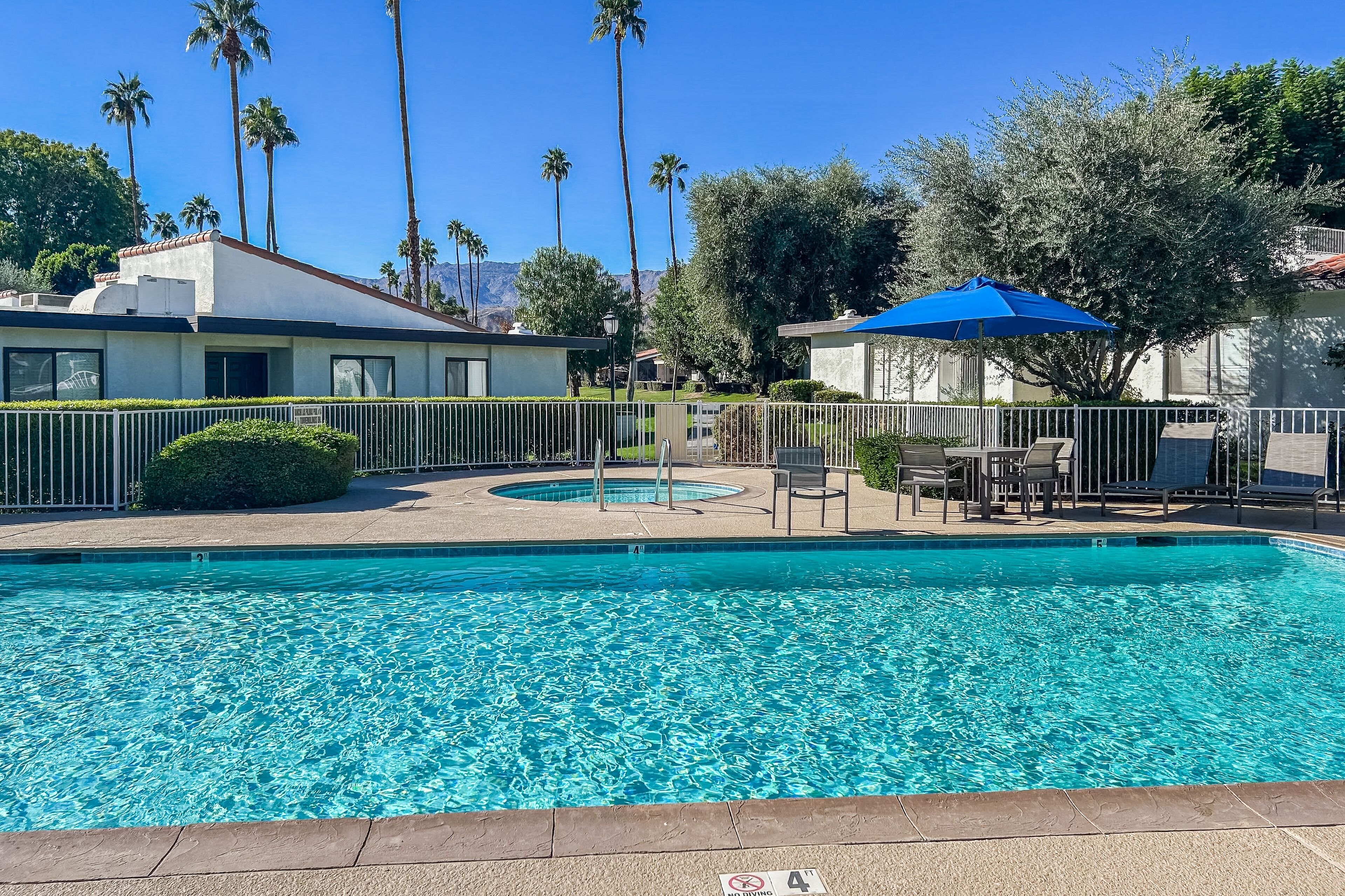 A swimming pool with a blue umbrella, surrounded by a fence, and palm trees in the background.