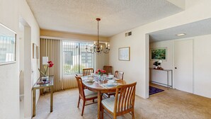 Un comedor con una mesa redonda, sillas y una araña. Hay una ventana con cortinas y una puerta que da a otra habitación.