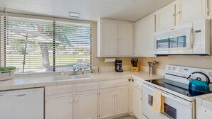 Una cocina con armarios blancos, microondas, estufa y ventana con persianas.