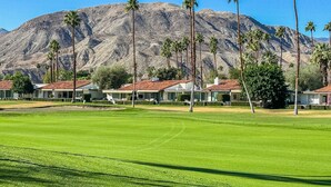 Un campo de golf con montañas al fondo, palmeras y una hilera de casas con techos de tejas rojas.