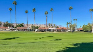 Un campo de golf con cielo despejado, palmeras y edificios con tejados rojos al fondo.