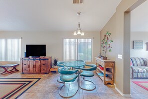 A spacious living room with a large flat-screen TV, a dining area with a glass-top table and chairs, and a cozy seating area with a patterned sofa.