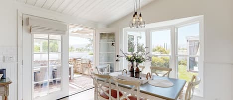 A bright dining area with white wooden furniture, a chandelier, and large windows offering views of the outdoors.
