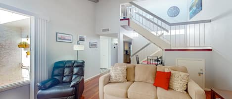 A spacious living area with a leather armchair, a beige sofa with patterned cushions, a wooden floor, a staircase with a railing, and a wall clock.