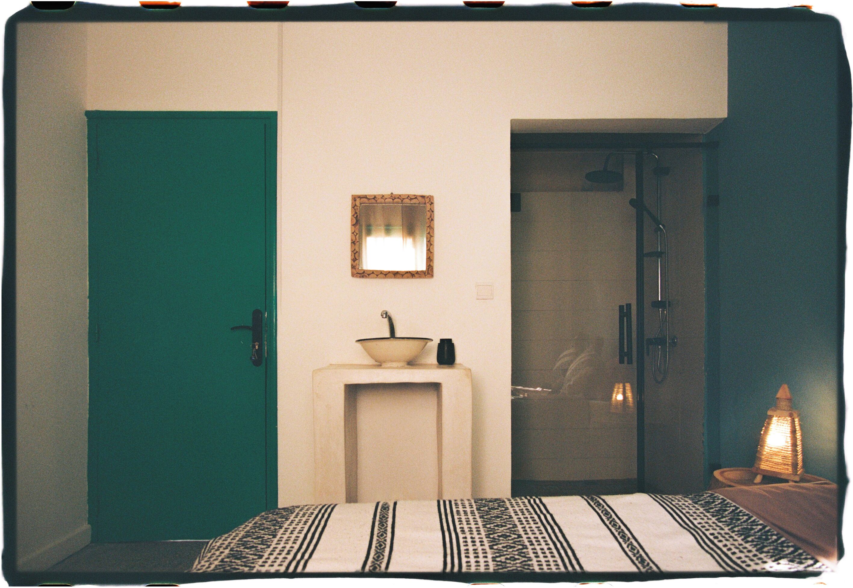 Une pièce avec une porte de couleur turquoise, un petit lavabo avec un receveur, un miroir, une douche et un lit avec une literie à motifs.
