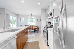 A modern kitchen with a stainless steel refrigerator, white cabinetry, and a dining area with wooden chairs and a chandelier. - Dreamy, dog-friendly home w/ private pool, spa & terrace - near El Paseo (Palm Desert)