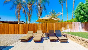 Une terrasse avec quatre chaises longues et une table placée sous un grand parasol, le tout entouré d’une clôture en bois et de palmiers.