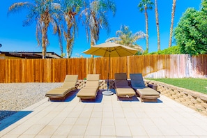 A patio with four lounge chairs and a table under a large umbrella, surrounded by a wooden fence and palm trees. - Dreamy, dog-friendly home w/ private pool, spa & terrace - near El Paseo (Palm Desert)