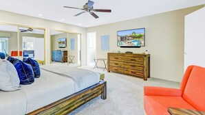 Een slaapkamer met een bed, een plafondventilator, een televisie, een houten dressoir en een fel oranje stoel.