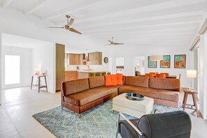 A spacious living room with a large brown sectional sofa, a white ottoman, and a blue patterned rug. There are ceiling fans, a kitchen area in the background, and artwork on the walls. - Racquet Club East Home with Modern Flair | Pool, Spa & Outdoor Living (Palm Springs)