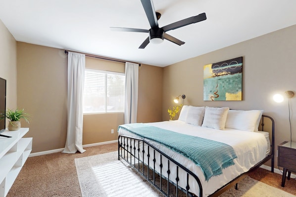 Une chambre à coucher comprenant un lit, un ventilateur de plafond, des œuvres d’art, une lampe de chevet et une fenêtre avec des rideaux.