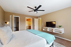 Une chambre à coucher avec un lit, un ventilateur de plafond, un téléviseur posé sur une étagère blanche et une porte donnant sur une autre pièce.