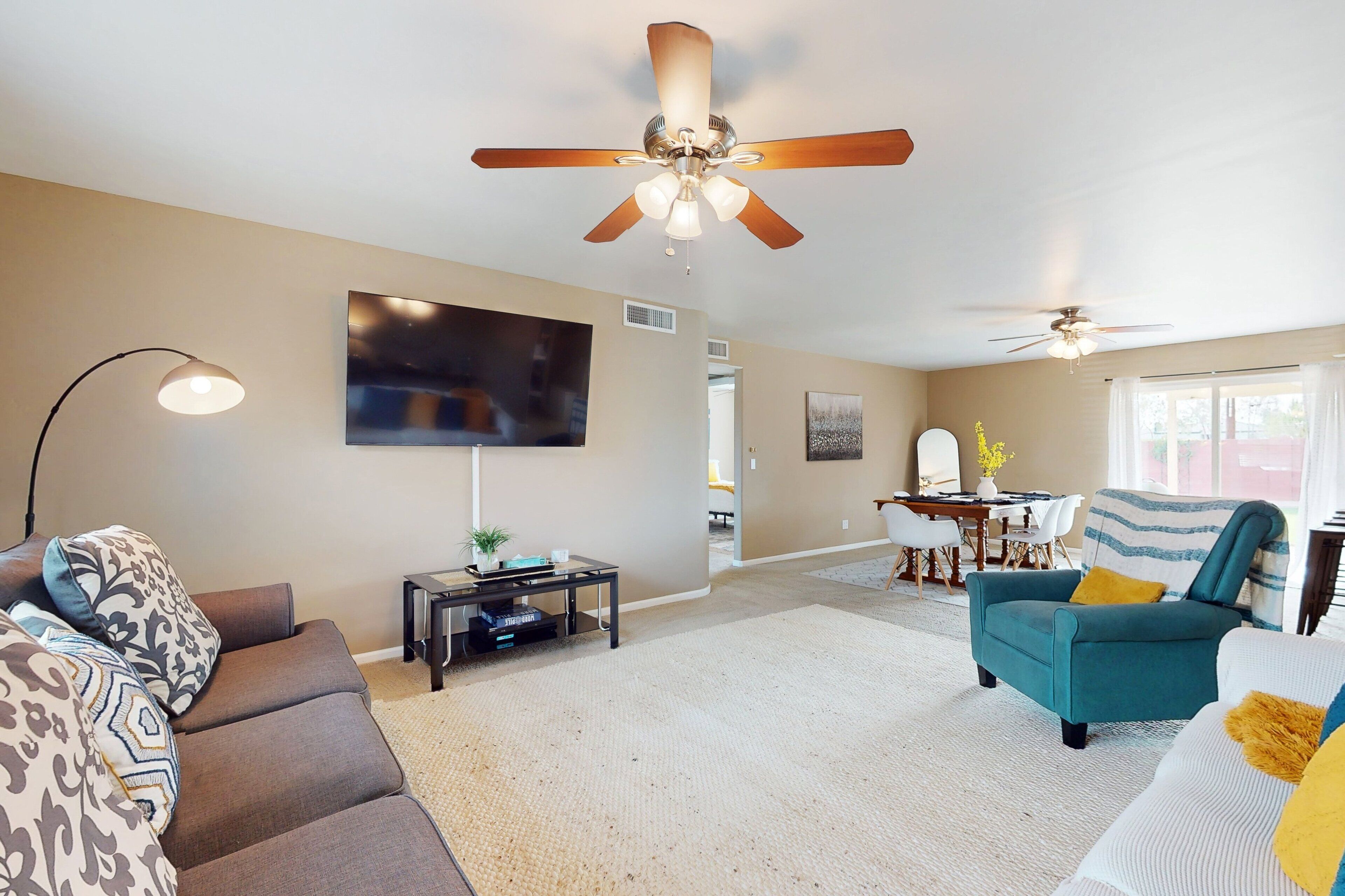 A living room with a sofa, armchair, and a coffee table. There is a ceiling fan, a flat-screen TV mounted on the wall, and a dining area with a table and chairs.