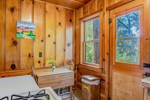 Interior de una cabaña de madera con una mesa de madera, una ventana, un bote de basura y un cuadro en la pared.