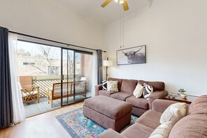 Ein Wohnzimmer mit einer Sofagarnitur, einem Couchtisch und einer Stehlampe. Ein Glastür öffnet sich zu einem Außenbereich mit einem Holztisch und Stühlen.