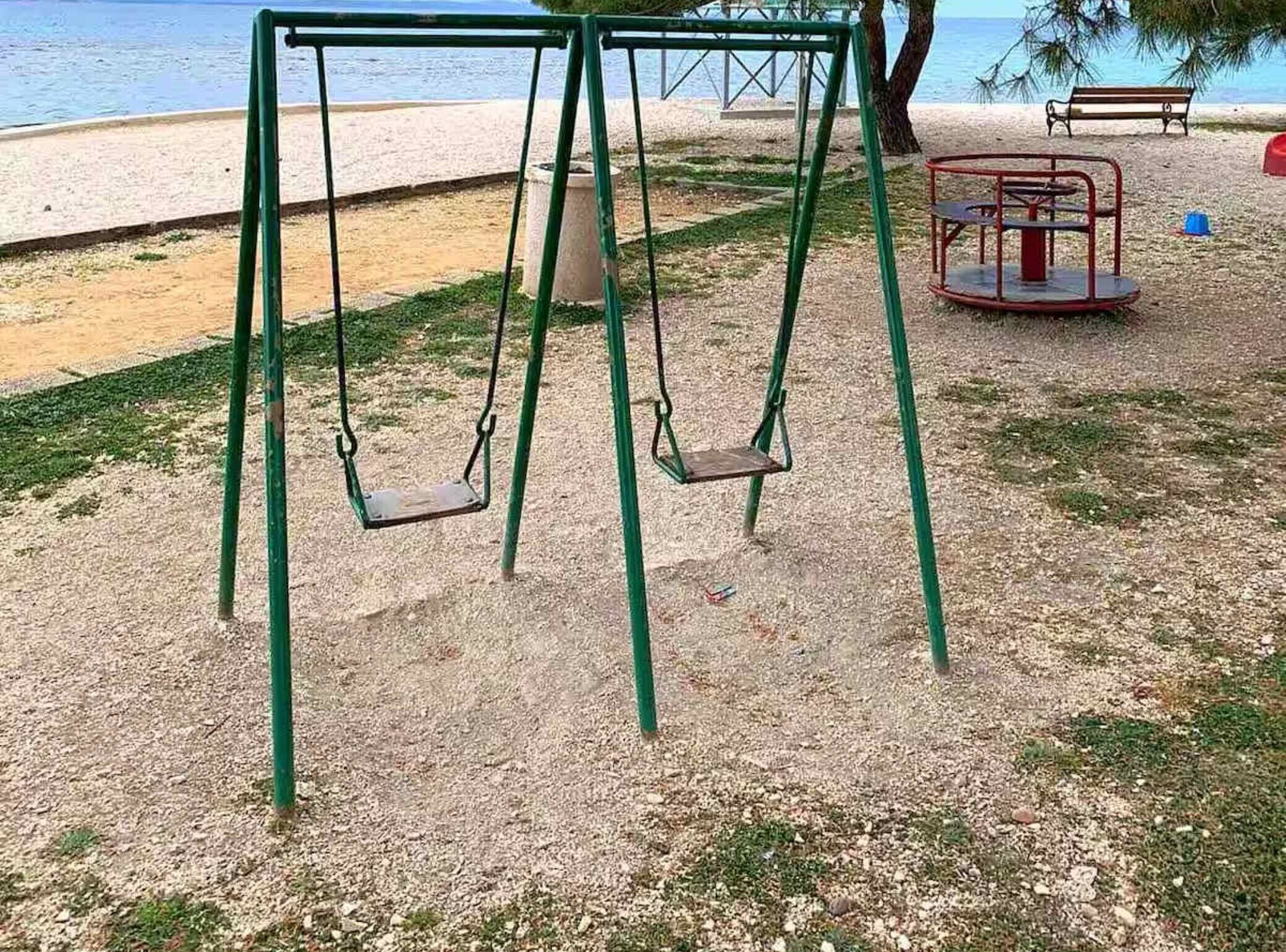 Un ensemble de balançoires dans un parc, avec un sol sablonneux, un banc et une vue sur la mer.