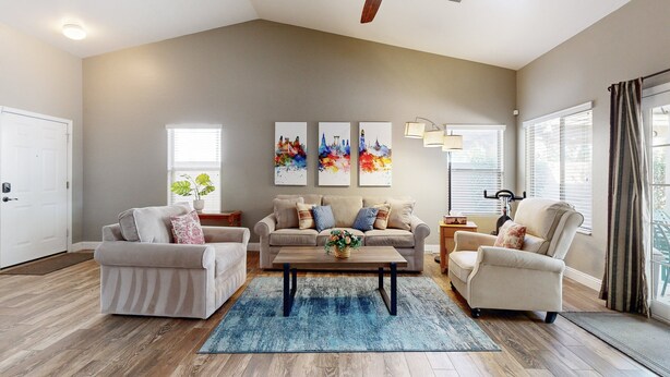 A living room with a beige sofa, two armchairs, a coffee table, and a ceiling fan. There are three paintings on the wall and a window with blinds. - Modern home with large yard, mountain views, & convenient location (Phoenix)