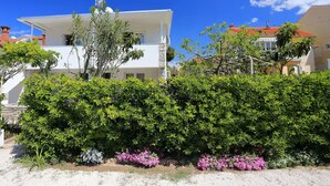 Een wit, twee verdiepingen hoog huis met een rood dak, omringd door weelderige groene struiken en roze bloemen.