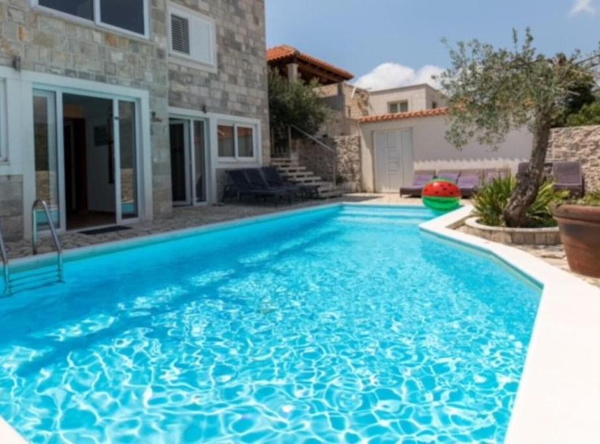 Una piscina con agua azul cristalina, rodeada de edificios de piedra y una pequeña zona ajardinada con una decoración de sandía.