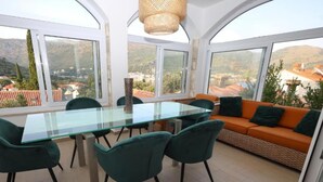 Ein modernes Zimmer mit einem Glastisch und Stühlen, einer Couch und großen Fenstern mit Blick auf eine Stadt und Hügel.