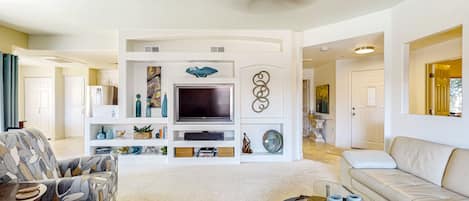 A modern living room with a large flat-screen TV, a sofa, a coffee table, and decorative items on shelves.