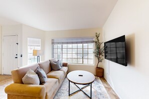 Un salon comprenant un canapé, une table basse ronde, un téléviseur fixé au mur et une fenêtre avec des stores.