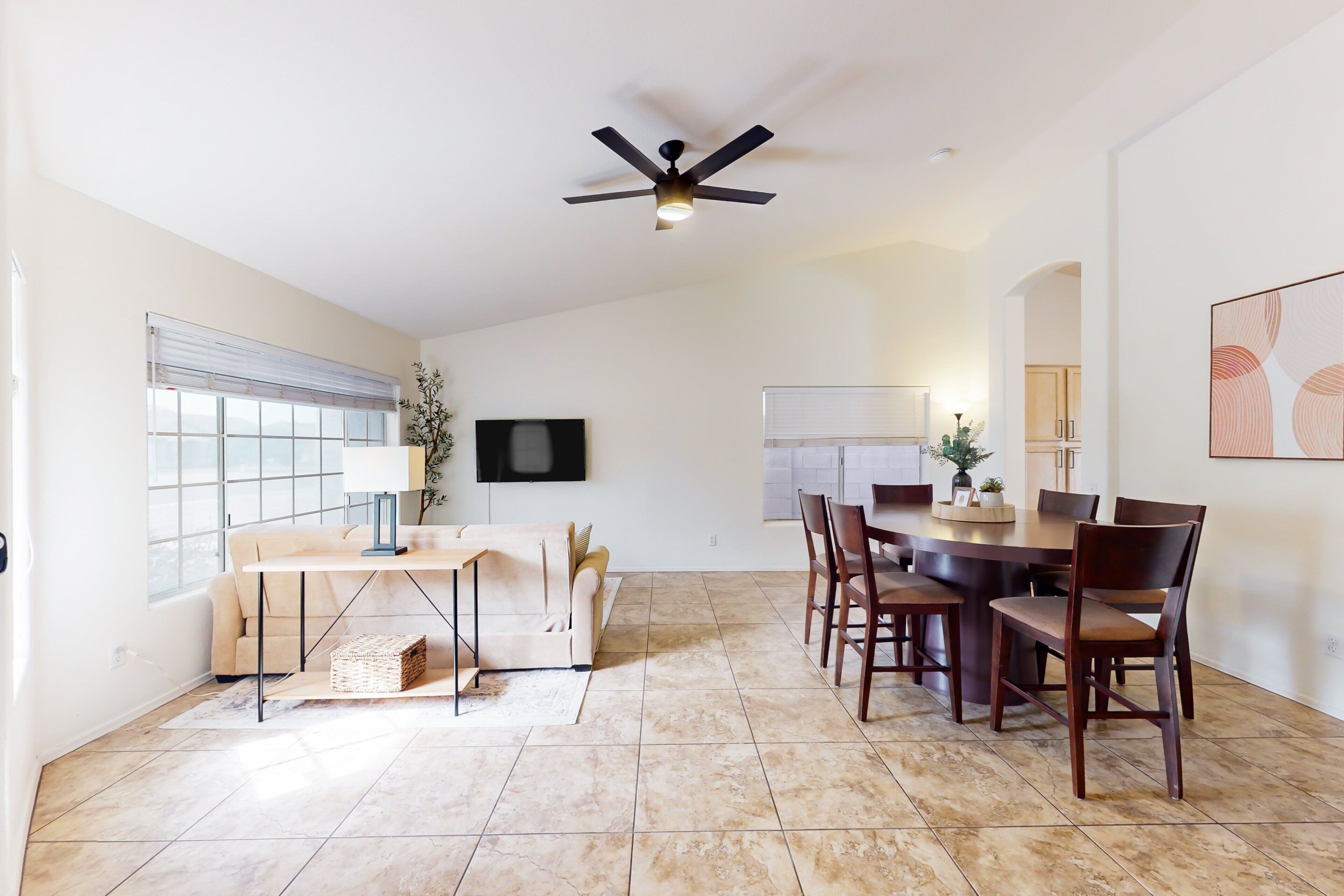 A spacious dining area with a high ceiling fan, a large window with blinds, and a view into the kitchen.