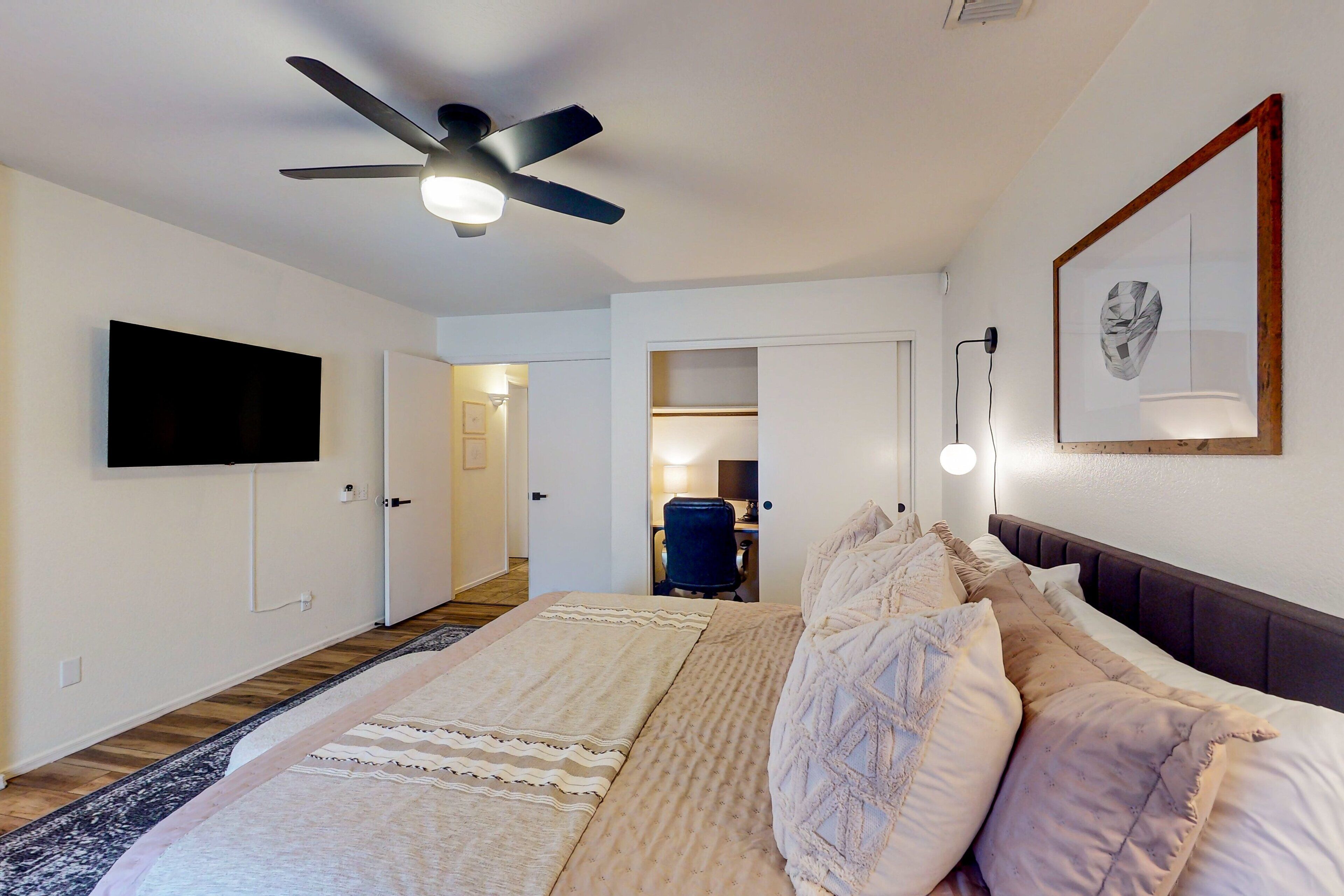 A bedroom with a bed, a ceiling fan, a flat-screen TV, a framed picture, and a desk with a chair.
