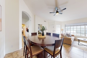 A spacious dining area with a round table and chairs, a ceiling fan, and a large window providing natural light.