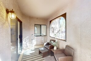 Ein sonnendurchflutetes Zimmer mit einem Korbsessel, einem kleinen Tisch, einer Topfpflanze und einem Fenster mit Jalousien.