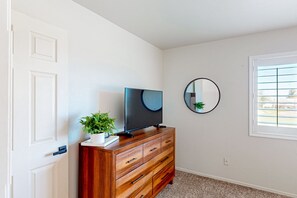 A room with a wooden dresser, a flat-screen TV, a potted plant, a round mirror, and a window with blinds. - Golf-course-front home with private pool, spa, terrace & mountain view (Oro Valley)