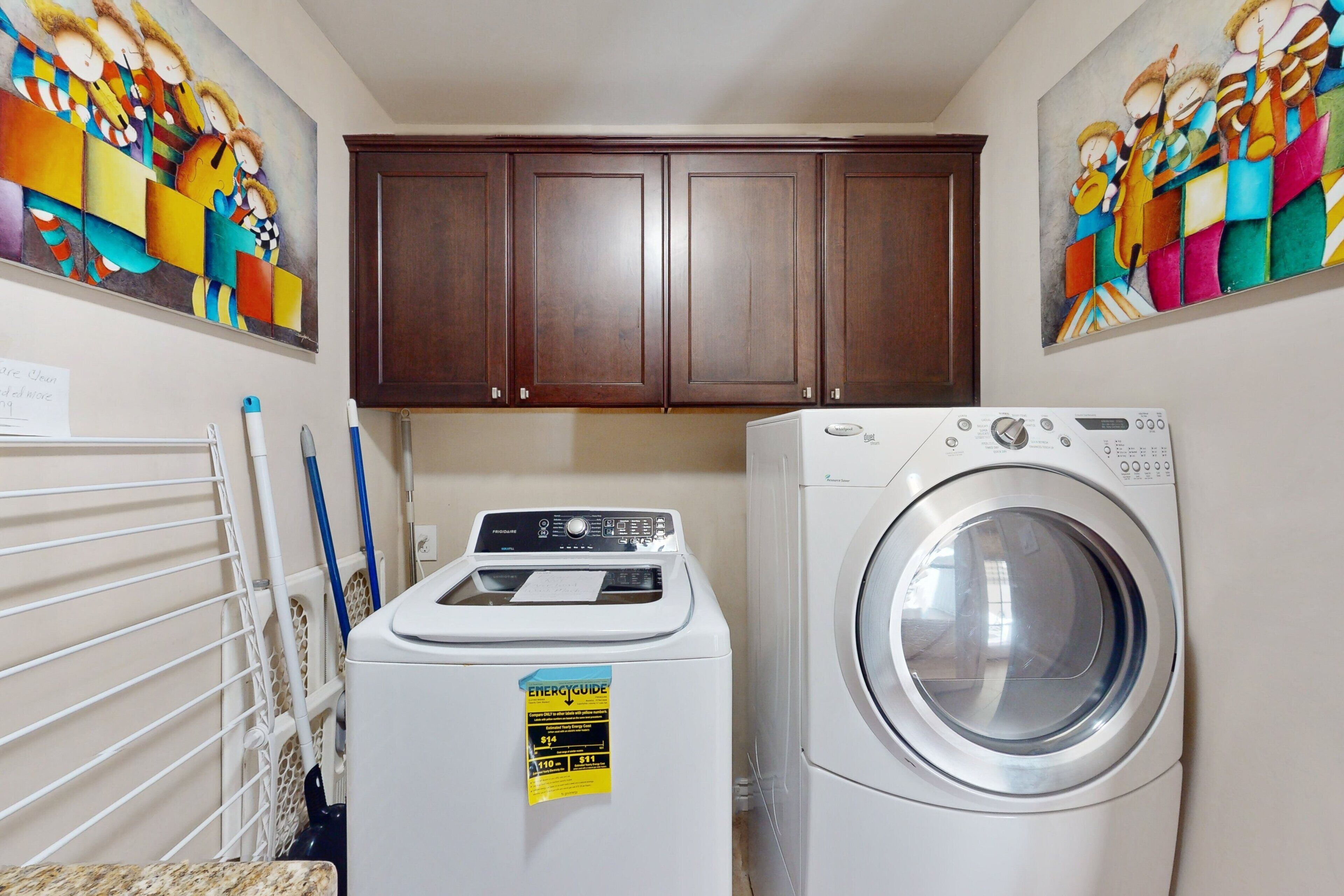 A laundry room with a front-loading washer and dryer, wooden cabinets above, and colorful abstract art on the walls.