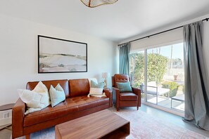 A modern living room with a brown leather sofa, a wooden coffee table, and a brown armchair. There is a large window with curtains, a framed abstract painting on the wall, and a view of the outdoors. - Fully remodeled condo near Fashion Square with pool, fireplace, patio, streaming (Scottsdale)