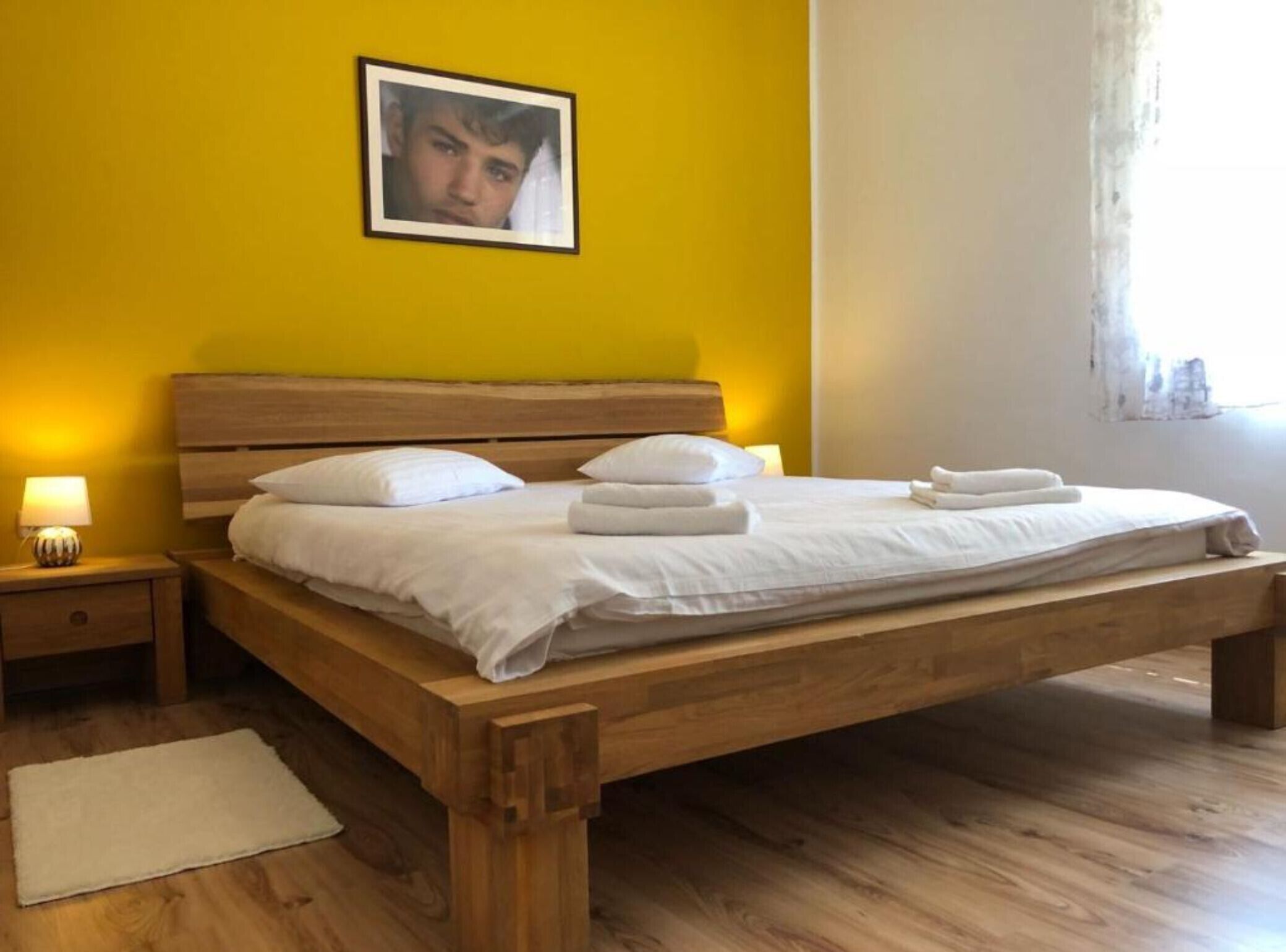 A wooden bed with white bedding and towels, a bedside lamp, and a framed picture on a yellow wall.