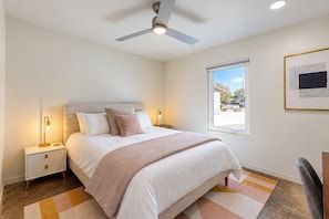 Ein Schlafzimmer mit Bett, Nachttischen, Deckenventilator, Fenster und einem gerahmten Bild an der Wand.