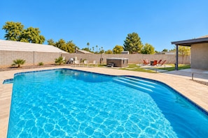 A rectangular swimming pool with clear blue water, surrounded by a tiled deck. There are lounge chairs and a barbecue area in the background.