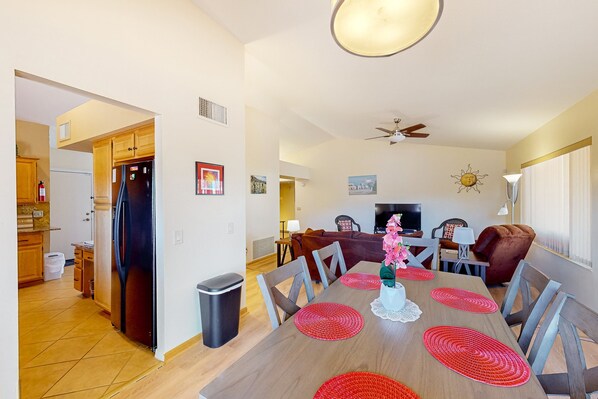 A dining area with a wooden table, red placemats, and chairs. There is a ceiling fan, a TV, and a fireplace in the background. - Mountain-view home with shaded patio, large yard, on-site golf & tennis (Tucson)