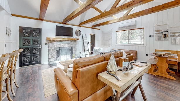 A spacious living area with a high ceiling, exposed wooden beams, a stone fireplace, leather seating, and a dining table with a decorative centerpiece.
