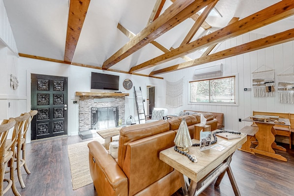 A spacious living area with a high ceiling, exposed wooden beams, a stone fireplace, leather seating, and a dining table with a decorative centerpiece. - Dog-friendly, renovated home with fireplace & amazing deck (Idyllwild)