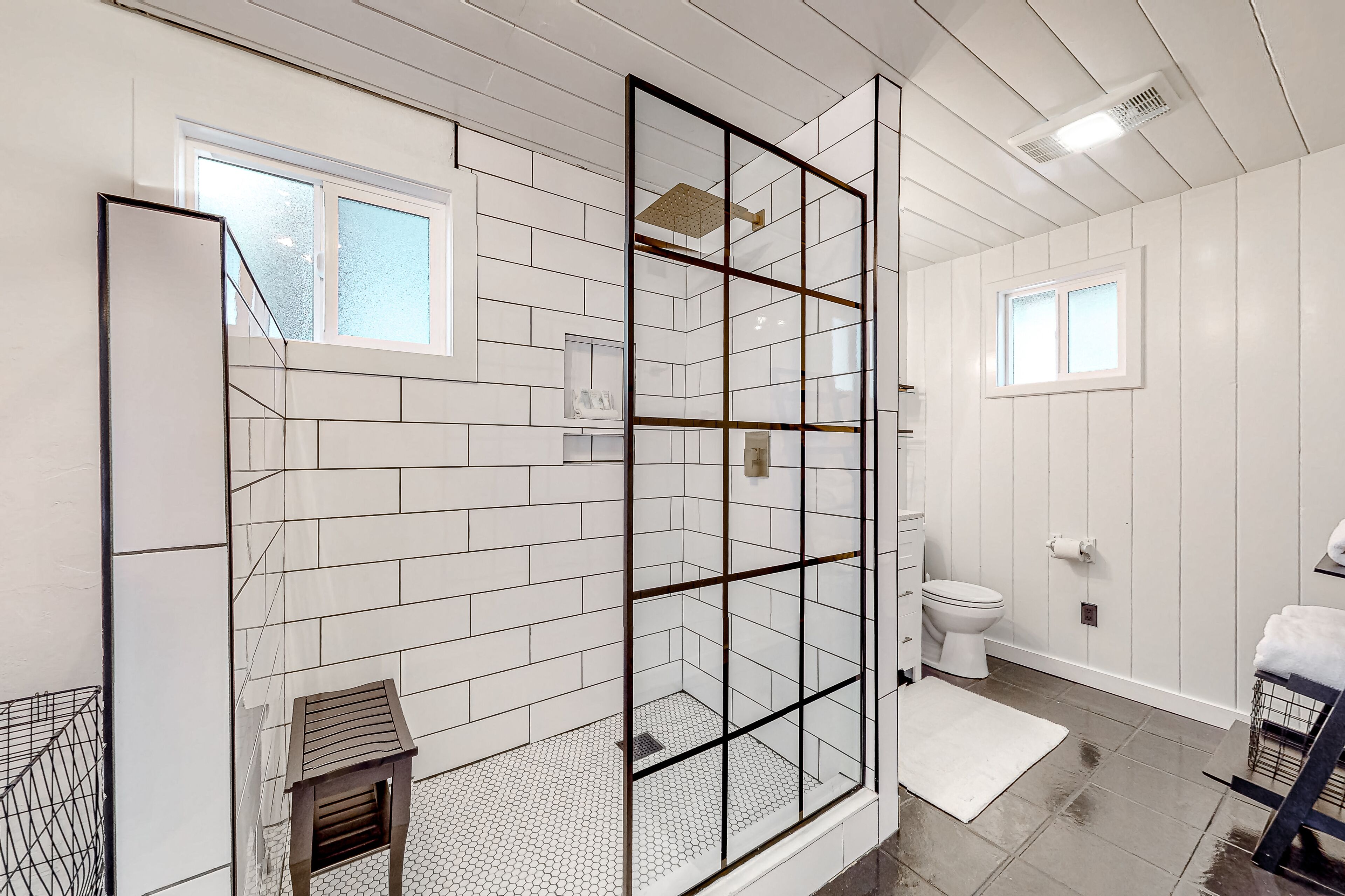 Un baño moderno con paredes de azulejos blancos, cabina de ducha con mampara de vidrio, inodoro y un pequeño taburete de madera.